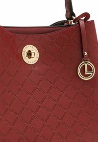 Rote Handtasche mit geprägtem Logomuster, goldfarbener Hardware und einem abnehmbaren runden Anhänger. Glatte Textur und eine strukturierte Form.