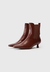Par de botas de tobillo de cuero marrón con punteras puntiagudas, paneles laterales elásticos y tacones cortos en forma de kitten sobre un fondo blanco.