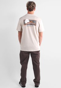 T-shirt en coton beige avec un design graphique représentant des montagnes et un soleil au dos ; associé à un pantalon marron et des chaussures noires.