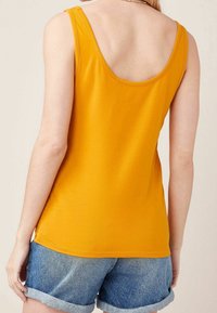 Débardeur jaune moutarde avec un large dos échancré, en tissu doux, associé à un short en denim bleu clair retroussé.