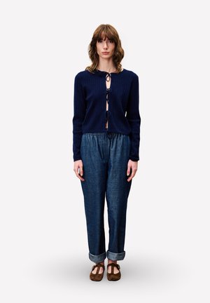 Femme portant un cardigan bleu marine à nouer sur le devant, un pantalon en denim ample avec des poignets, et des chaussures Mary Jane marron, se tenant devant un fond blanc uni.