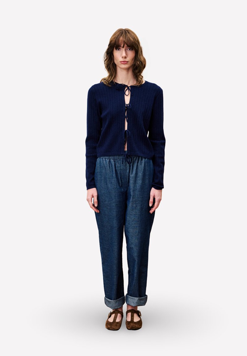 Vrouw in een marineblauwe cardigan met strik, loszittende denim broek met omslag en bruine Mary Jane-schoenen, staande tegen een eenvoudige witte achtergrond.