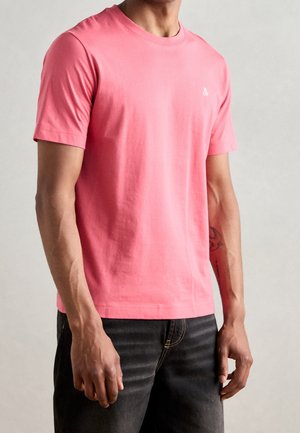 Mann trägt ein einfaches korallenrosa kurzärmeliges T-Shirt und dunkle Jeans, steht vor neutralem Hintergrund.