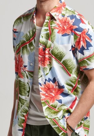 Uomo con camicia a maniche corte sbottonata con grandi fiori rossi e foglie verdi sopra una maglietta bianca, mano in tasca.