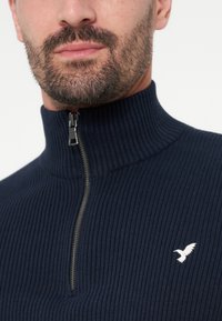 Homme barbu portant un pull zippé côtelé bleu marine avec un col haut et un petit logo blanc d’oiseau sur la poitrine.