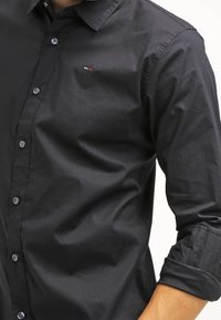 Camisa negra de botones confeccionada en tejido suave, con un cuello clásico, mangas largas y un pequeño logo cerca del pecho.
