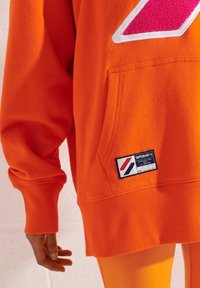 Sudadera de algodón naranja con un bolsillo tipo canguro, que presenta un logo en rosa y blanco y una etiqueta negra con acentos en rojo y azul.