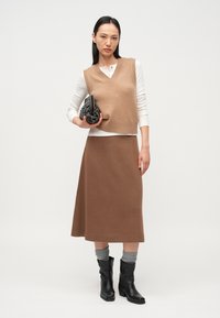 Beige gebreid vest over een wit shirt met lange mouwen, gecombineerd met een bruine rok tot op de knie. Zwarte leren enkellaarzen en een zwarte handtas met textuur.