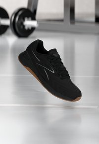 Μαύρο αθλητικό παπούτσι Reebok με σόλα καουτσούκ που αιωρείται πάνω από ανακλαστικό δάπεδο γυμναστηρίου, βάρη και μεταλλικός εξοπλισμός γυμναστηρίου θολωμένοι στο φόντο.