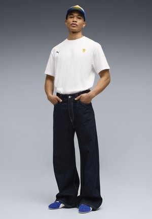 Młody mężczyzna stoi z rękami w kieszeniach, ubrany w białą koszulkę Puma Ferrari, szerokie ciemne dżinsy, niebieskie sneakersy i niebiesko-żółtą czapkę na szarym tle.