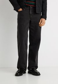 Άνδρας που φοράει μαύρα τζιν με ίσια γραμμή, μαύρα μοκασίνια, ριγέ πουκάμισο και μαύρο σακάκι με το ένα χέρι στην τσέπη μπροστά σε λευκό φόντο.