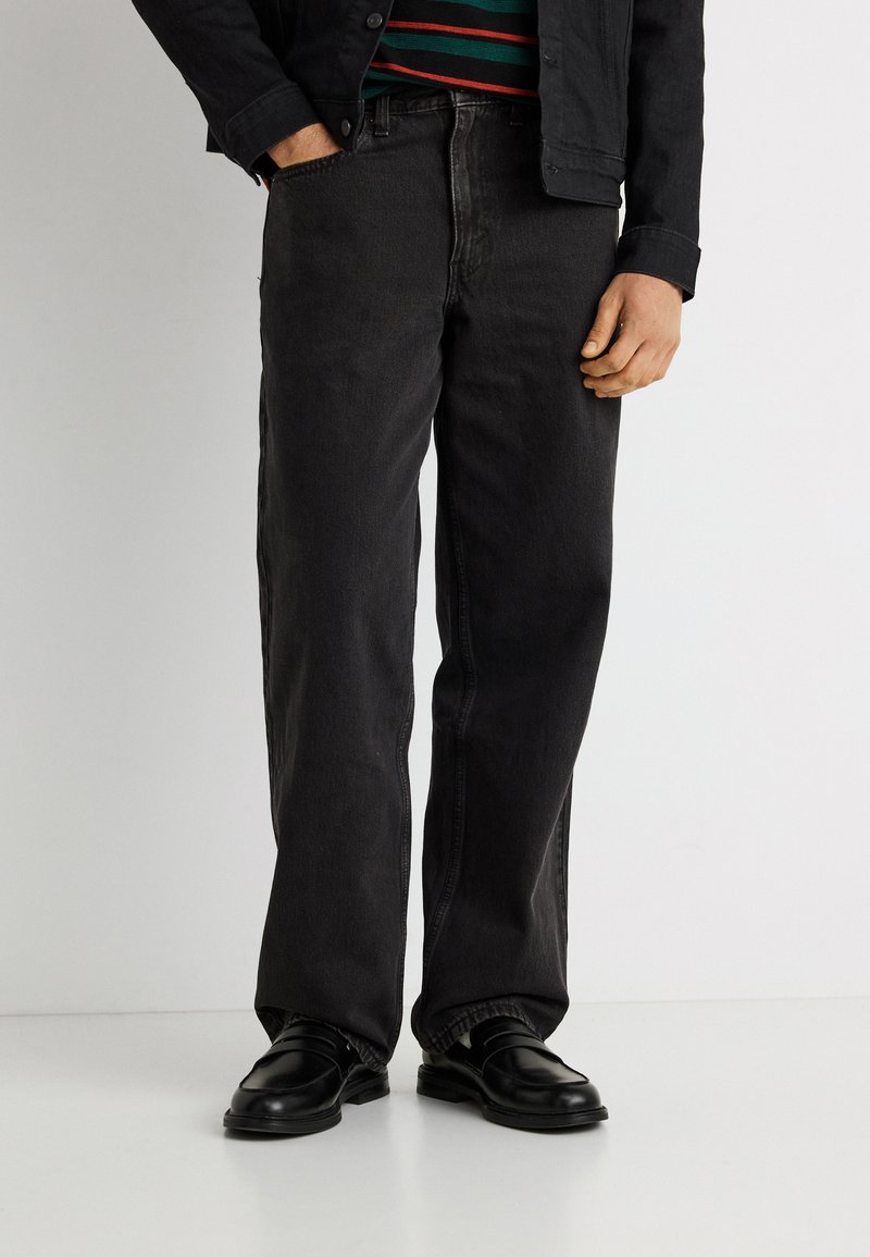 Άνδρας που φοράει μαύρα τζιν με ίσια γραμμή, μαύρα μοκασίνια, ριγέ πουκάμισο και μαύρο σακάκι με το ένα χέρι στην τσέπη μπροστά σε λευκό φόντο.