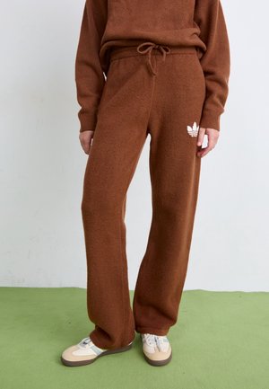 Persoon draagt bruine gebreide broek met Adidas-logo, bijpassende trui en witte sneakers, staand op een groene vloer tegen een witte muur.