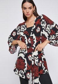 Top de estilo kimono en negro, rojo y blanco con un patrón floral; presenta mangas amplias y una cinturilla con lazo. Tejido ligero con un diseño fluido.