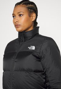 Black puffer jacket with a high collar, featuring horizontal quilting and a white logo on the left chest. Made of glossy, synthetic material.