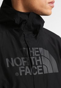 Schwarze wasserdichte Jacke mit hohem Kragen und übergroßem Logodruck. Glatte Textur mit schlankem Schnitt, aus leichtem Material gefertigt.