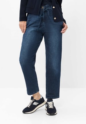 Frau trägt dunkelblaue High-Waist-Jeans mit Bindegürtel und schwarze Sneakers mit weißen Schnürsenkeln und silbernen Akzenten, steht auf weißem Boden.