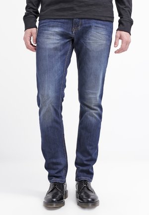 Mann trägt schmal geschnittene blaue Jeans mit ausgewaschenen Details und schwarze Lederschuhe, steht vor einem weißen Hintergrund.