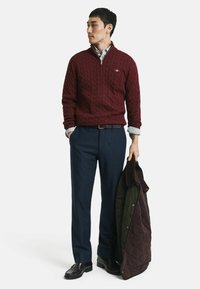 Burgunderfarbener Kabelstrickpullover mit einem Viertel-Reißverschluss, kombiniert mit marineblauen Hosen und schwarzen Schuhen, hält eine dunkelgrüne und braune gesteppte Jacke.