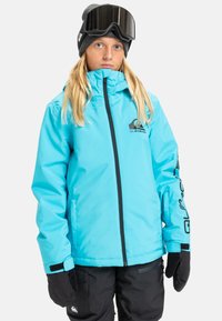 Veste imperméable bleue avec fermeture éclair à l'avant, accents noirs, logo sur la poitrine et poignets ajustables. Associée à un pantalon de neige sombre et des gants noirs.