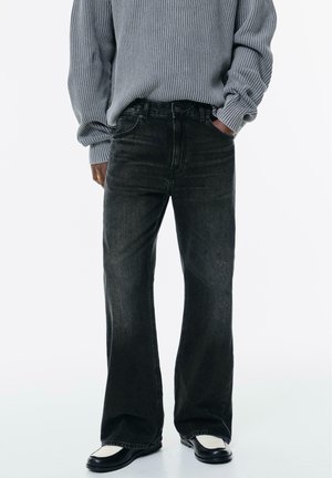 Personne portant un jean noir à jambes larges, un pull gris côtelé et des chaussures noir et blanc, debout avec une main dans la poche.