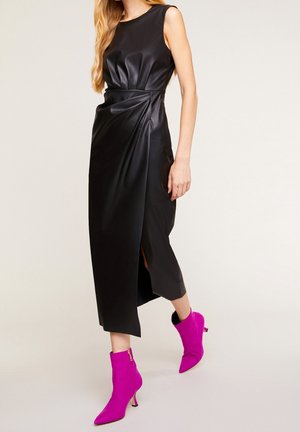 Femme portant une robe midi en cuir noire sans manches avec une fente à l'avant et des bottines à talons aiguilles roses vives, debout sur un fond clair.