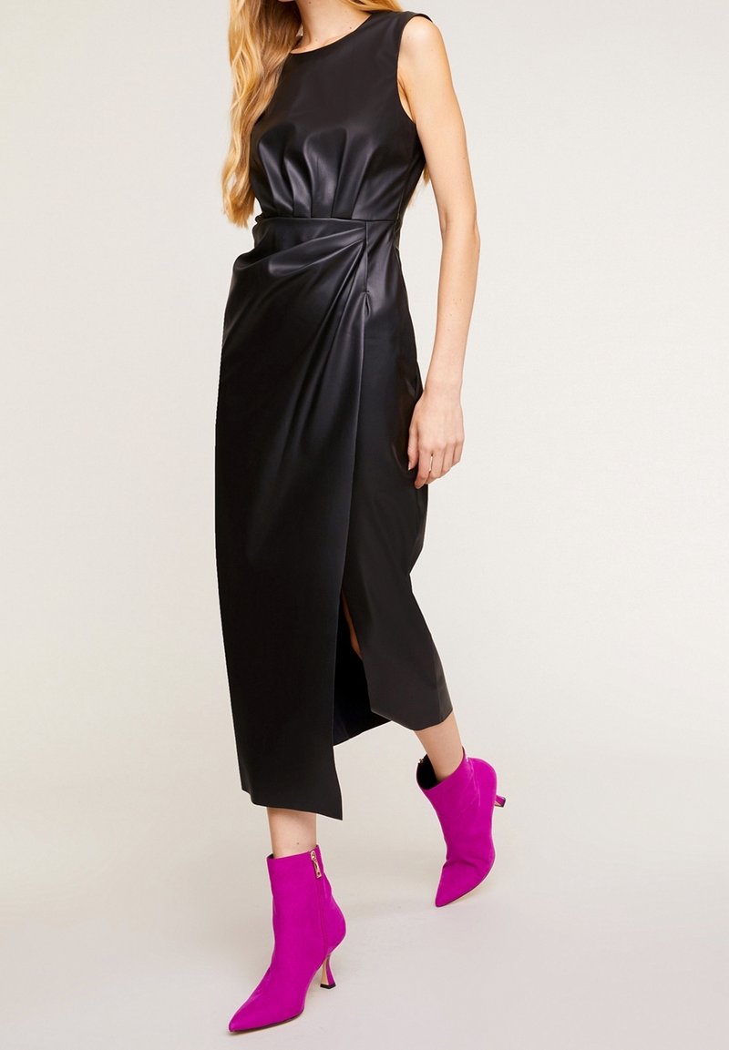 Femme portant une robe midi en cuir noire sans manches avec une fente à l'avant et des bottines à talons aiguilles roses vives, debout sur un fond clair.
