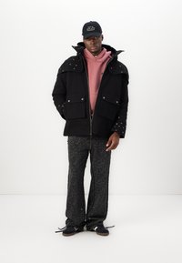 Chaqe oscuro exterior con acentos de tachuelas, sudadera rosa debajo, pantalones oscuros con estampado y una gorra negra con texto blanco.