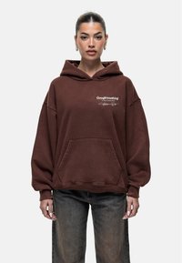 Felpa oversize marrone con tasca frontale e testo del logo. Il tessuto sembra morbido; presenta un cappuccio con cordoncino e maniche larghe.