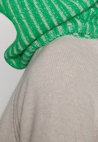 Grün-weiß gestreifter Strickpullover, der über einem hellbeigen, glatt strukturierten Pullover getragen wird. Sichtbare Nahtdetails auf dem beigen Pullover.