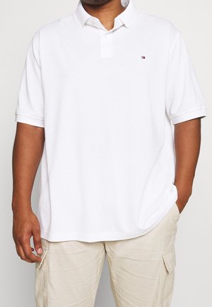 Mężczyzna w białej, krótkorękawowej koszulce polo z małym logo na piersi i jasnobeżowych spodniach bojówkach, z ręką w kieszeni.