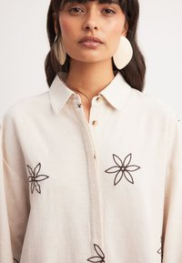 Ľanová košeľa v odtieňoch off-white, s hnedým kvetinovým vyšitím. Návrh s gombíkmi a límcom, s voľným strihom.