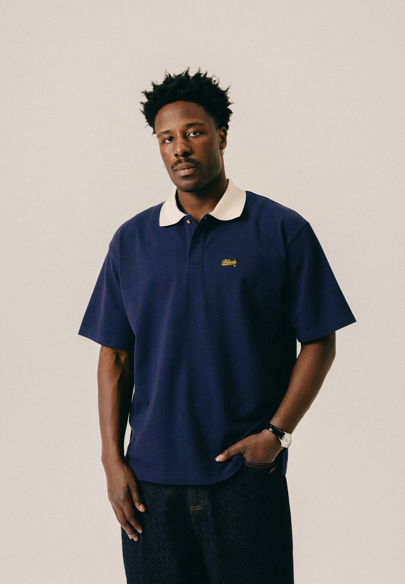 Homme aux cheveux courts et bouclés portant un polo bleu marine à col blanc, un jean foncé et une montre-bracelet, debout devant un fond clair.