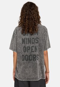 Camiseta gris de gran tamaño con un audaz texto negro en la parte de atrás que dice "MENTES ABIERTAS PUERTAS ABIERTAS". Tiene una textura desgastada y bordes sin acabar.