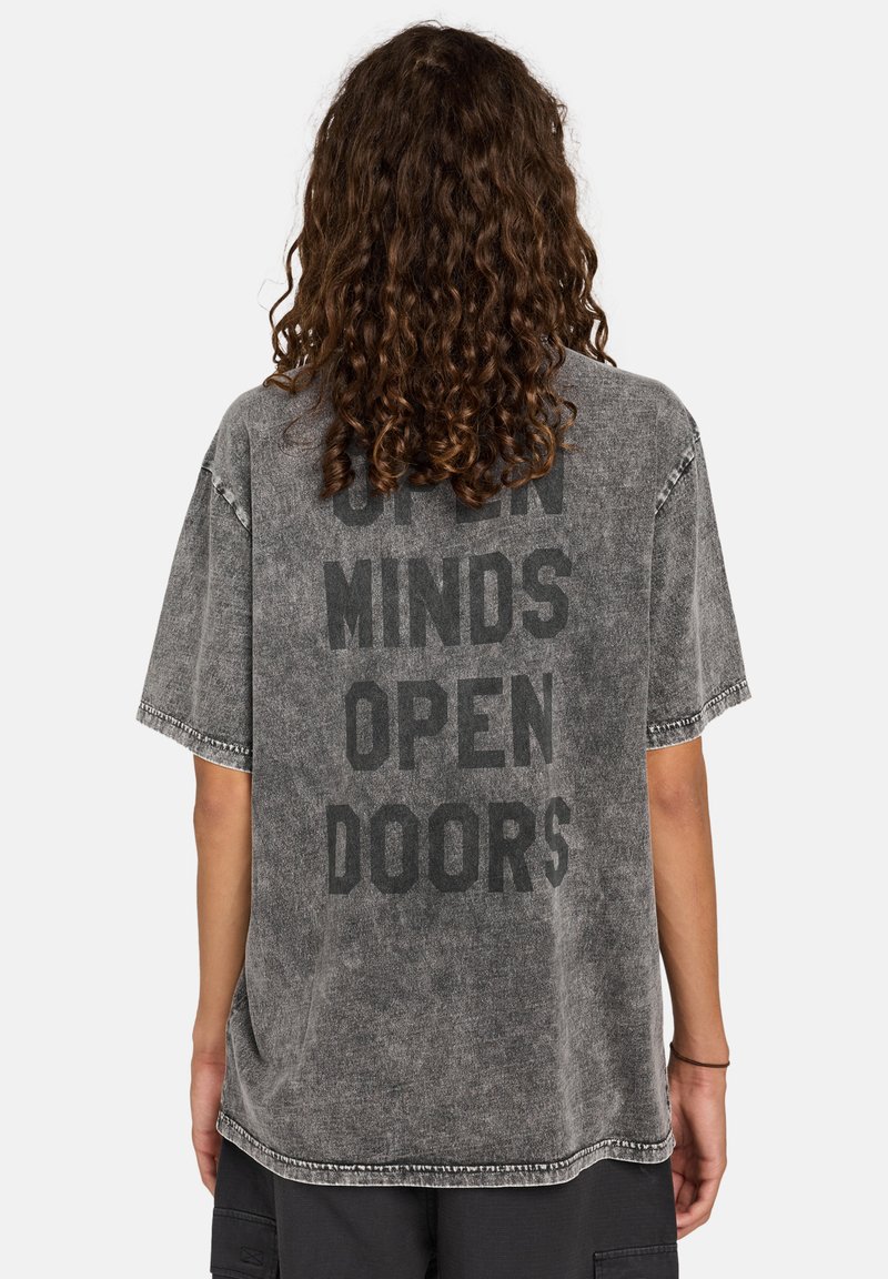 Camiseta gris de gran tamaño con un audaz texto negro en la parte de atrás que dice "MENTES ABIERTAS PUERTAS ABIERTAS". Tiene una textura desgastada y bordes sin acabar.