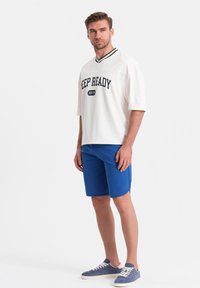 Bílá halenka s krátkým rukávem s nápisem "KEEP READY", modré šortky a modré tenisky. Texturovaná látka, sportovní design, uvolněný střih.