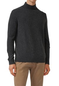 Uomo che indossa un maglione a collo alto a coste grigio scuro con maniche lunghe e pantaloni beige, in piedi con le braccia rilassate.