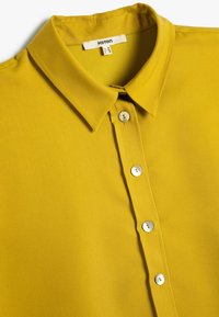 Camicia gialla senape con colletto, caratterizzata da una chiusura con bottoni frontale con cinque bottoni e una trama del tessuto liscia. Etichetta del marchio visibile.