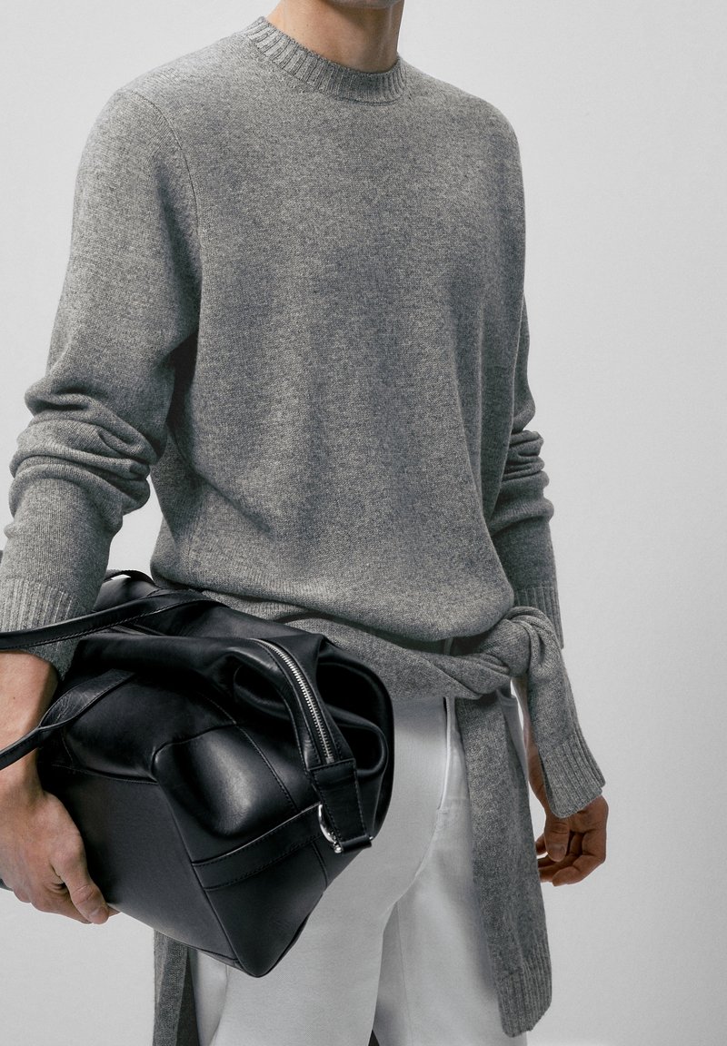 Grijze gebreide trui met geribde halslijn en manchetten, gecombineerd met een zwarte leren tas. De trui heeft een relaxed fit en lange mouwen.