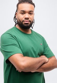 Άνδρας με γενειάδα και κοτσίδες dreadlocks φορώντας πράσινο μπλουζάκι στέκεται με σταυρωμένα τα χέρια μπροστά σε απλό λευκό φόντο.