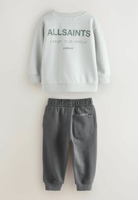 Harmaa huppari, jossa on "ALLSAINTS" -logo ja teksti vihreällä, yhdessä tummanharmaiden verkkareiden kanssa, joissa on logoprintti ja elastinen vyötärö.