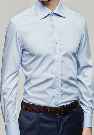 Chemise à manches longues bleu clair avec un col pointu et des boutons blancs, associée à une ceinture à motifs foncés et un pantalon navy.