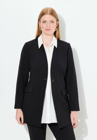 Blazer nero con chiusura a un solo bottone, due tasche frontali, vestibilità sartoriale, indossato sopra una camicia bianca con colletto, design minimalista, texture liscia.