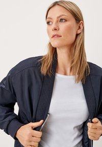 Vrouw met steil blond haar die een wit shirt en een marineblauwe jas draagt, de randen van de jas vasthoudend en naar de zijkant kijkend.