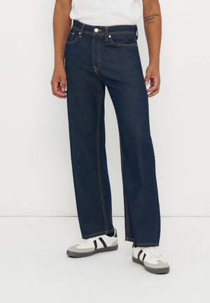 Un mannequin porte un jean bleu foncé à jambes larges avec des surpiqûres contrastantes, un haut blanc et des baskets blanches, sur un fond de studio uni.