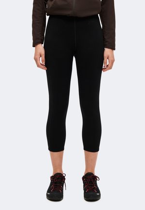 Personne portant un legging court noir, des chaussures de randonnée noires et rouges, et une veste marron, debout sur un fond clair uni.