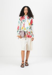 Camicia floreale con fiocco, con fiori rossi, gialli e blu su sfondo bianco; pantaloni corti crema; borsa a tracolla lavanda; sandali neri.