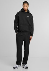 Hanorac negru cu buzunar frontal, având un logo alb. Asortat cu pantaloni negri și adidași alb-gri care oferă un contrast.