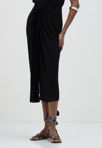Femme portant une jupe midi noire avec des détails drapés, des sandales à motifs enroulées autour des pieds, et un bracelet en argent sur un fond blanc uni.