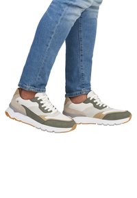 Les baskets présentent un mélange de daim et de textile crème, vert olive et taupe. Semelle blanche légère avec des accents texturés et de lacets épais.